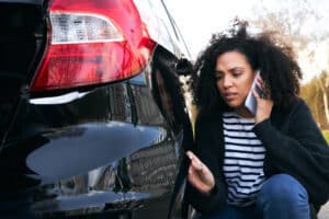 Mujer joven enfadada de pie junto a un coche vandalizado en un aparcamiento informando de los daños a la compañía de seguros mediante un teléfono móvil