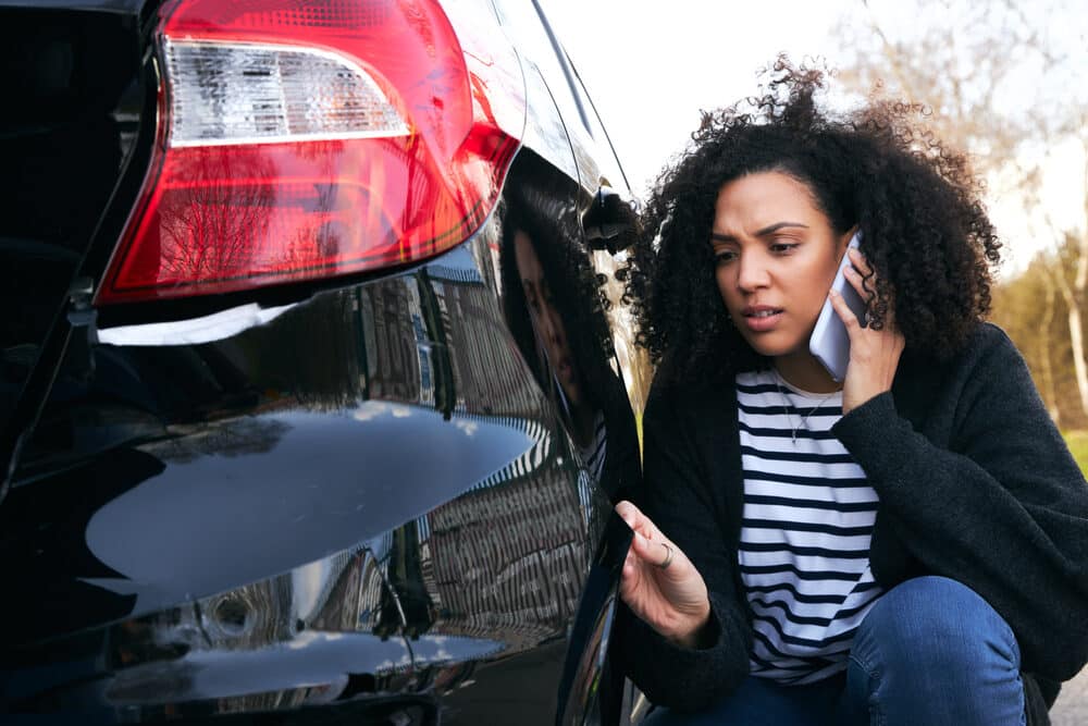 Mujer joven enfadada de pie junto a un coche vandalizado en un aparcamiento informando de los daños a la compañía de seguros mediante un teléfono móvil
