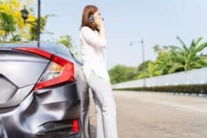 Mujer asiática molesta e infeliz que ha tenido un accidente de tráfico o ha chocado durante su viaje por carretera, conductora sentada junto al coche accidentado o averiado y usando el teléfono móvil para llamar al servicio de asistencia en carretera.