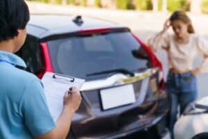 Agente de seguros rellenando el formulario de reclamación por accidente de vehículo en un portapapeles, perito de automóviles documentando los daños del vehículo mientras el conductor se siente afectado al fondo.