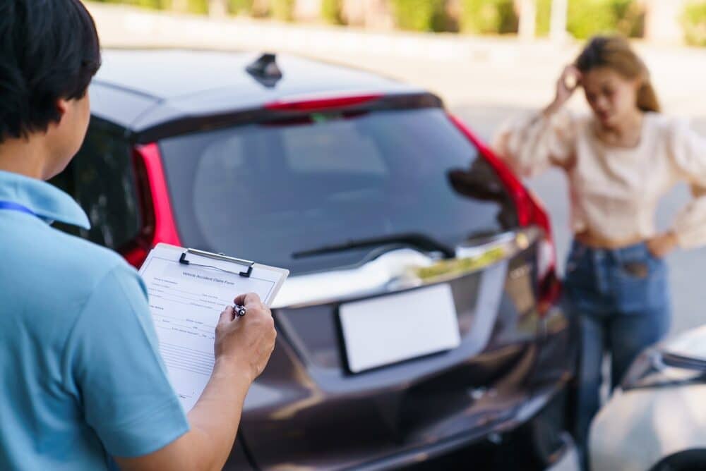 Agente de seguros rellenando el formulario de reclamación por accidente de vehículo en un portapapeles, perito de automóviles documentando los daños del vehículo mientras el conductor se siente afectado al fondo.