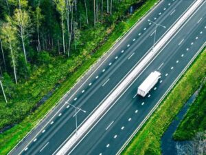 Vista aérea con dron de camiones semirremolques de transporte de mercancías y coches circulando por una carretera asfaltada a través de bosques verdes de verano.