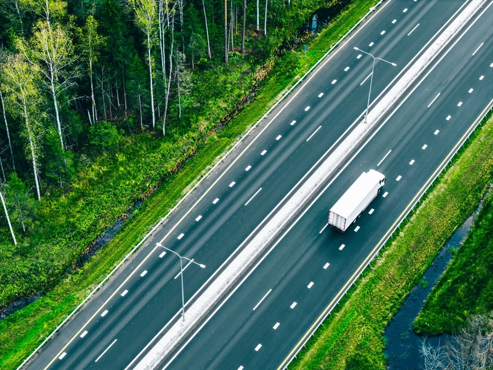 Vista aérea con dron de camiones semirremolques de transporte de mercancías y coches circulando por una carretera asfaltada a través de bosques verdes de verano.