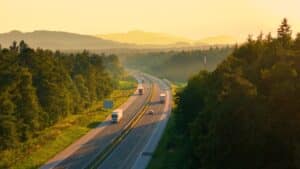 Una autopista atraviesa un hermoso paisaje boscoso moteado por una luz dorada. Camiones y coches circulan por la autopista cuando los primeros rayos de la mañana iluminan el campo cubierto de un frondoso bosque.